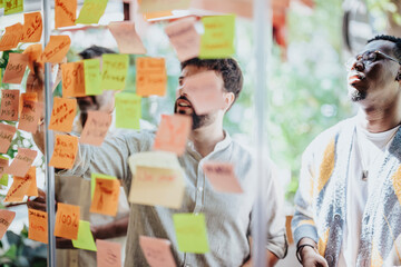 Diverse business partners collaborate in a modern office, brainstorming and planning with post-it notes on a glass wall. Successful teamwork and cooperation in achieving goals.