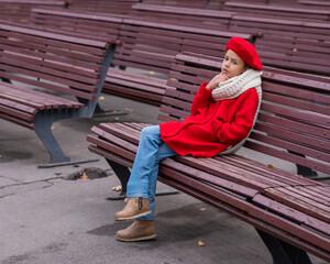 A sad Caucasian girl in a red coat and a beret sits alone on a bench. 