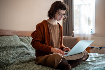 Busy thoughtful teenage girl sitting on bed with laptop computer, thinking, reading text document,...