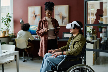 Young businessman in VR helmet sitting on wheelchair and preparing virtual presentation with female colleague using tablet - obrazy, fototapety, plakaty