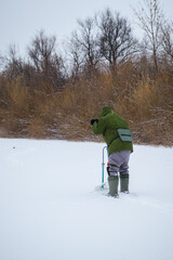 A fisherman is fishing in an ice hole on a frozen pond. The joy of winter fishing. A fisherman on a winter fishing trip on a cloudy day. 