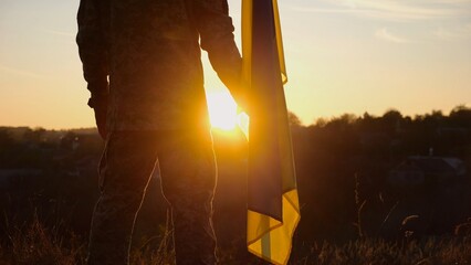 Male ukrainian army soldier stands with national banner against background of beautiful sunset....