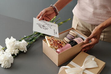 Woman putting handmade postcard with greetings for Mother Day into giftbox with white chocolate,...