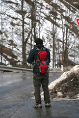 Fototapeta na wymiar A lone traveler with a red backpack stands on a winding road amidst a serene, snowy landscape, embodying solitude and adventure, concepts of journey, self-discovery, and nature’s majesty