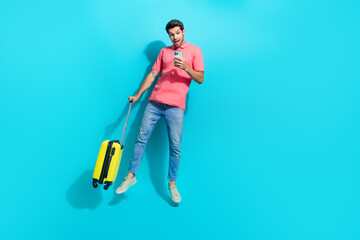 Full length photo of excited pretty man wear pink t-shirt jumping high holding baggage ordering taxi empty space isolated blue color background