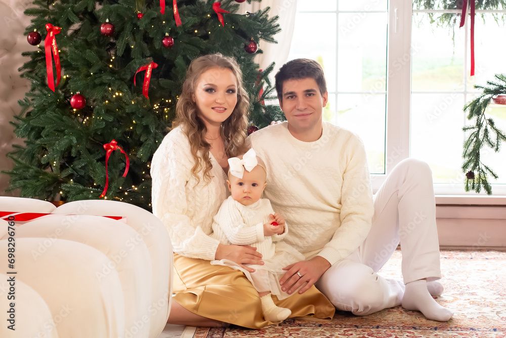 Wall mural a beautiful family with a little daughter in their arms sits on the floor near a beautiful christmas