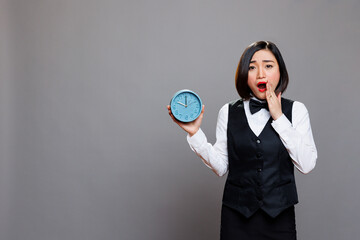 Hotel asian woman receptionist yawning while holding alarm clock and looking at camera in studio. Young attractive tired sleepy waitress oversleeping and showing time portrait