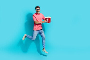Full length photo of cute excited man wear pink t-shirt jumping eating pop corn watching 3d movie empty space isolated blue color background