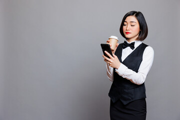 Young asian waitress wearing uniform drinking coffee to go and scrolling social networks on...