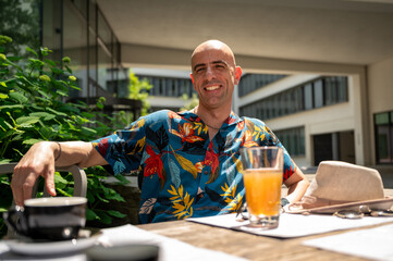Fashionable mid-age man carrying a colorful hawaiian shirt sits at a bar table with drinks and...