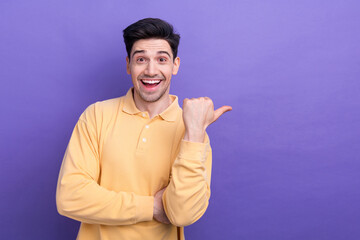 Photo of charming impressed man dressed yellow shirt pointing thumb empty space isolated purple...