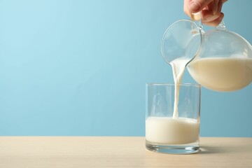 Woman pouring milk into glass at wooden table against light blue background, closeup. Space for text