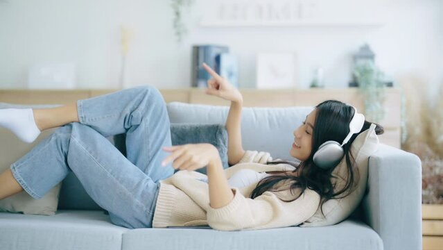 Young asian woman listening to music on couch in living room at home. Happy asia female using mobile smartphone, wearing headset and lying down on sofa
