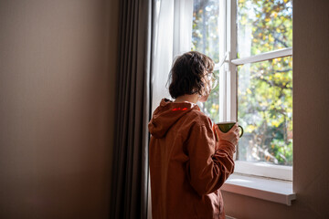 Lonely teenager standing near window, looking at sunny summer weather with sadness, sorrow, spending time alone at home. Unsociable teen girl having no friends. Youthful crisis, communication problems - obrazy, fototapety, plakaty