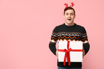Handsome young man in warm sweater and reindeer headband holding gift box on pink background