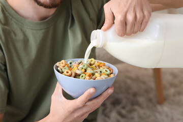 Man pouring milk into bowl with cereal rings in living room, closeup