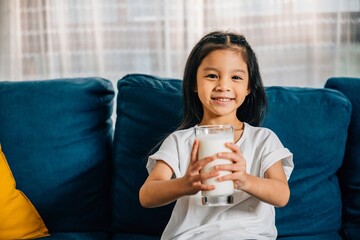 Close-up of a little Asian girl's hand holding a glass of milk while sitting on the sofa at home. This charming image depicts the importance of health care nutrition and the joy of childhood.