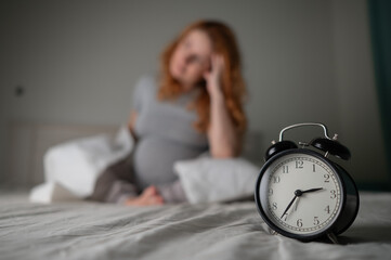 Caucasian pregnant woman sits on the bed and suffers from insomnia. Alarm clock in the foreground. 
