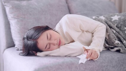 Asian woman feeling a headache and body warmth, anxious about getting sick or unwell while lying on her home sofa and wrapped in a blanket. Capturing the moment of discomfort and concern at home - Powered by Adobe