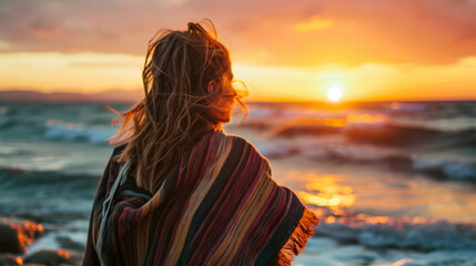 Woman looking at winter sunset on the beach with a shawl on her shoulders on a cold evening - obrazy, fototapety, plakaty