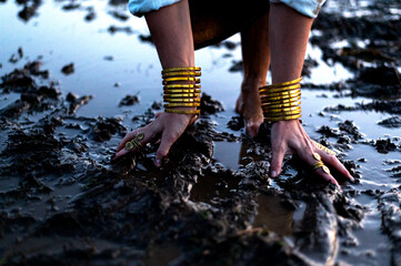 A woman's bare feet and hands with luxury bracelets and rings in a dirty puddle of black mud