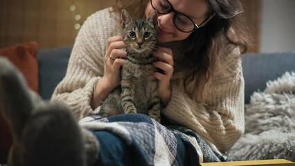 A young Caucasian woman petting her cute grey cat while sitting on the couch in the living room on a winter evening