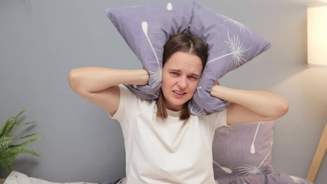 Angry sad unhappy Caucasian woman closing her ears with pillow in morning because of neighbor noise sleepless girl annoyed by alarm clock or loud neighbors