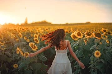 Tuinposter Carefree Happy beautiful young woman in white dress opened arms up in air and looking at sunset in a large field of sunflowers, Freedom concept, Enjoyment, Summer time. © oatawa