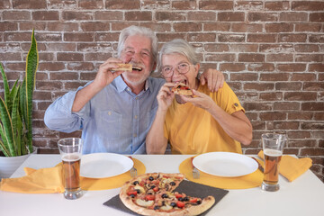 Happy senior couple looking at camera biting a slice of pizza. Elderly woman and man share a...