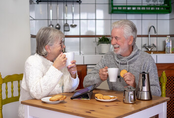 Portrait of attractive smiling senior couple having breakfast together sitting at kitchen table. Elderly 70s husband and wife enjoying relaxed retirement