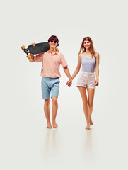 Fototapeta premium Summer vibe. Smiling happy young couple, man and woman holding hand, walking with skateboard against white background. Concept of summer vacation, travelling, retro style, fashion
