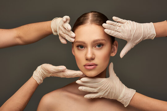 estheticians in medical gloves examining face of young woman on grey background, facial treatment