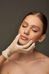 esthetician in medical glove touching face of pretty woman on grey background, dermatology concept