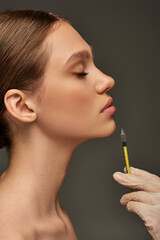 Obraz premium esthetician in medical glove holding syringe near lips of young woman on grey background, side view