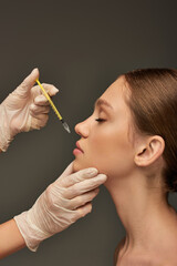 Obraz premium esthetician in medical glove holding syringe near lips of young woman on grey background, filler