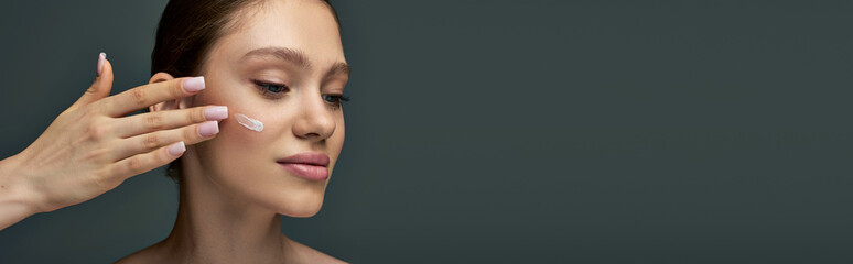 beautiful young woman with bare shoulders applying cream on face on grey background, banner