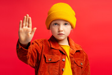 Serious little girl wearing yellow hat, showing stop gesture with hand isolated on red background