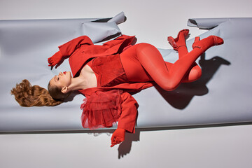 top view of young model in red outfit with tights and high heels lying on grey background, fashion