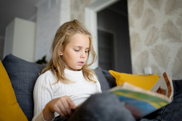A little girl with thick blond hair is immersed in the fascinating world of a book on a comfortable sofa.