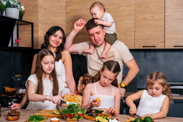 Large family - mom, dad, three daughters and baby son together prepare salad for lunch in modern...