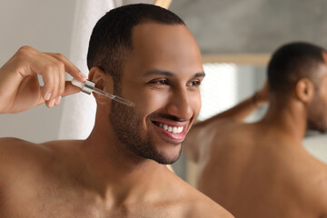Handsome man applying cosmetic serum onto face in bathroom, space for text