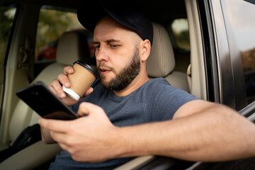 Male bearded driver with a smartphone in hand and paper cup of hot coffee. Concept of inattention at the wheel, rest and coffee break - obrazy, fototapety, plakaty