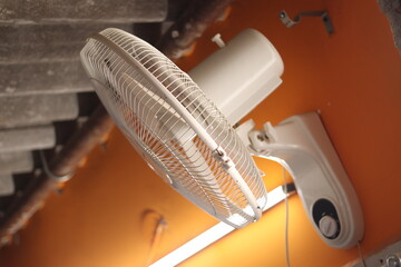 A white colored Wall revolving fan or table fan on an orange wall with tube light in background,...