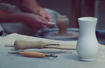Fototapeta na wymiar Workshop. Art Studio. Potter at work. White Clay Vase on Table. Potter's tools. Potter's hands on a Background. Filtered Photo