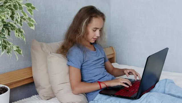A teenage girl with an open laptop is sitting on her bed typing in her bedroom