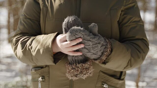 Woman in warm clothes takes off her knitted mittens in sunny winter park closeup. Warming of air with arrival of spring during change of seasons. Their relief from overly hot clothes on snowy morning.