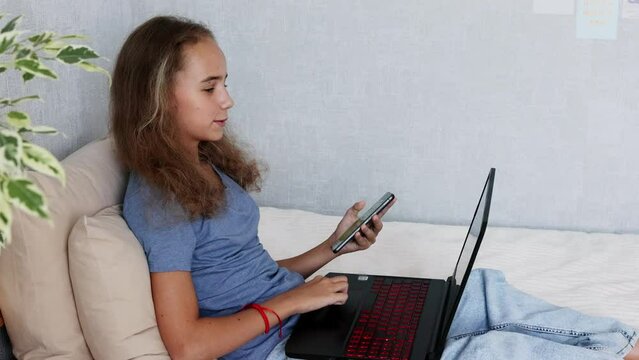 In the bedroom, a lovely teenage girl with blonde hair is sitting on the bed and typing on a laptop