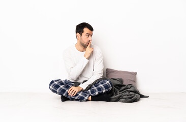Caucasian man in pajamas sitting on the floor at indoors showing a sign of silence gesture putting...