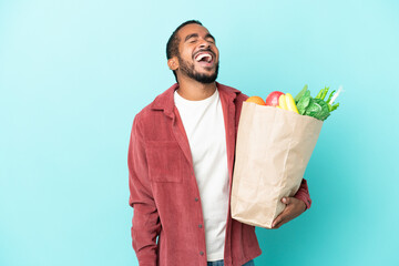 Young latin man holding a grocery shopping bag isolated on blue background laughing