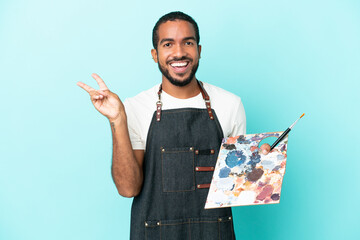 Young artist latin man holding a palette isolated on blue background smiling and showing victory...